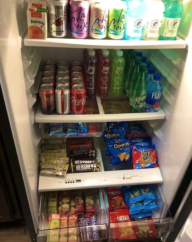 An open refrigerator filled with neatly organized drinks and snacks, including La Croix, Gatorade, Coca-Cola, Doritos…