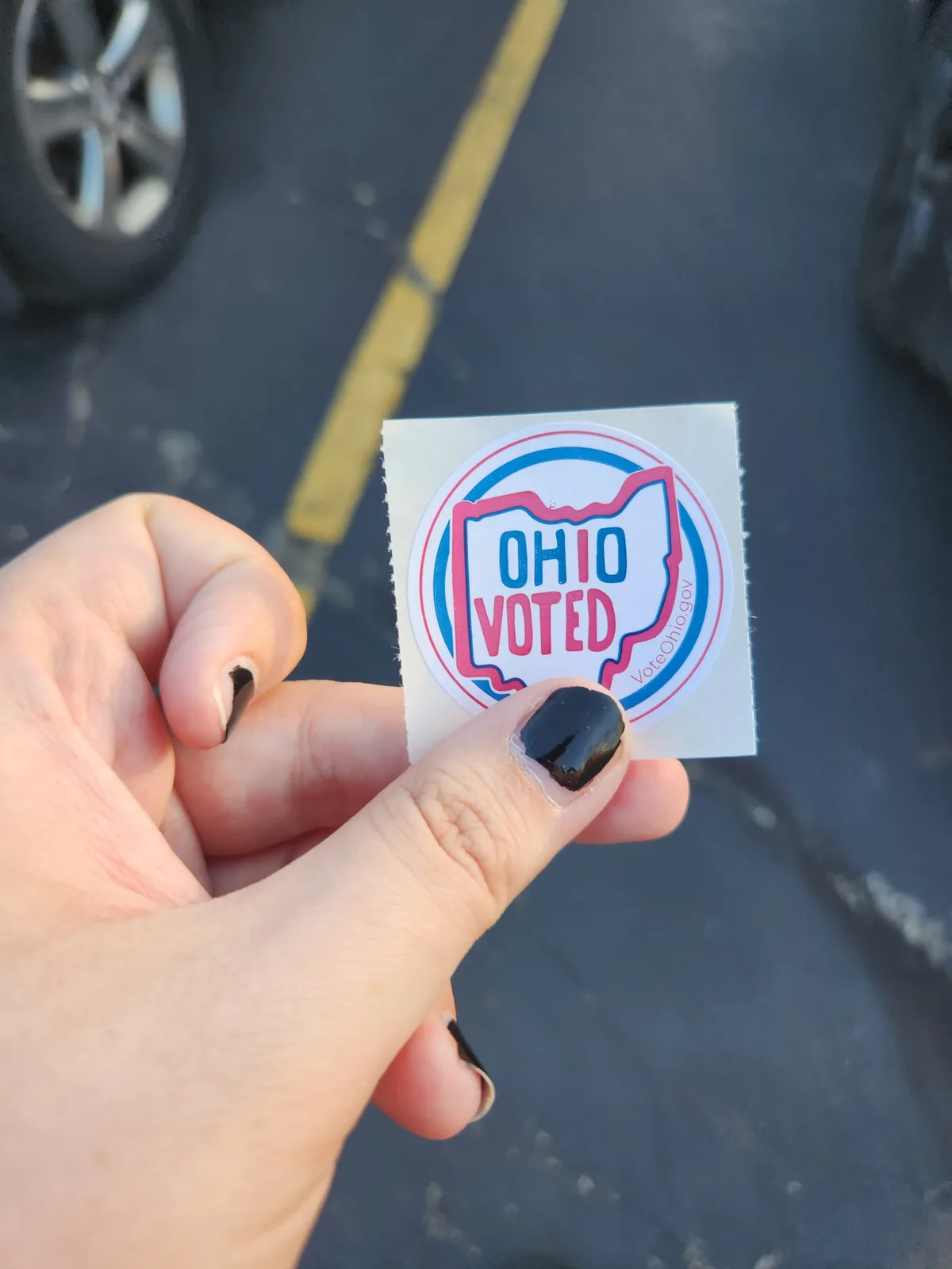 A hand with black nail polish holds up a circular 'OHIO VOTED' sticker featuring the state outline in pink and blue, with…