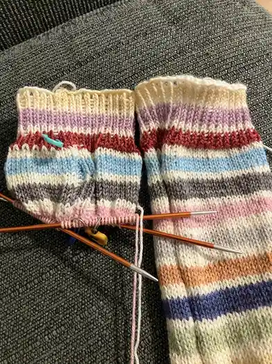 Two mismatched, colorful striped socks are shown side-by-side on a gray textured couch, one sock is fully knitted while the…