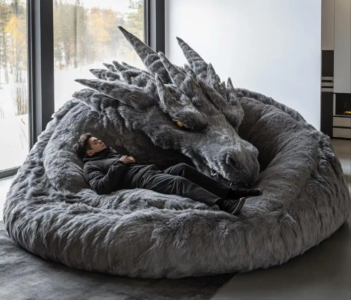 A person is lying down inside a giant, plush, grey dragon-shaped beanbag chair that curls around them like a cozy nest. The…