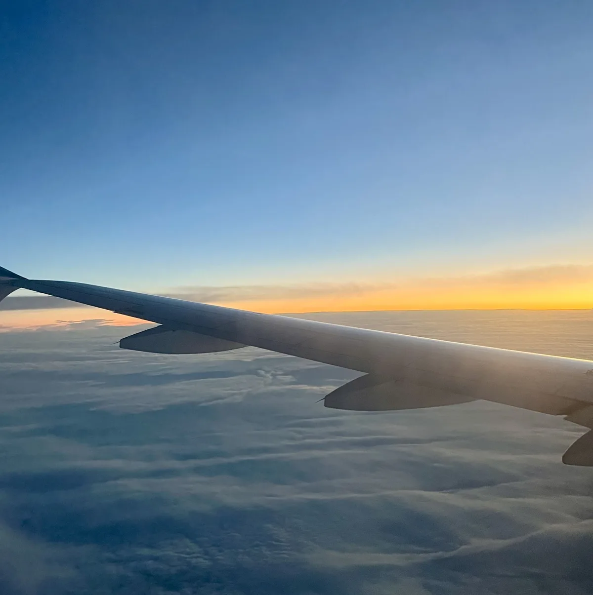 A serene view from an airplane window showing the wing against a gradient sky transitioning from blue to orange at the…