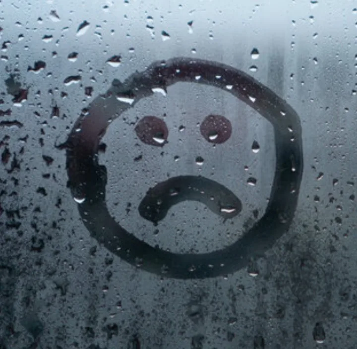 A sad face drawn in dark liquid on a wet, rain-speckled window, with the word 'sad' overlaid as text. The face has two round…