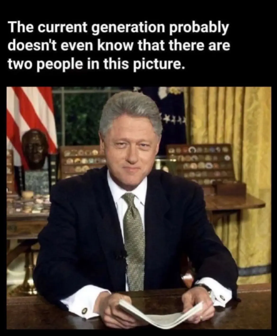 A photo of Bill Clinton sitting at a desk in the Oval Office, holding a newspaper, with an American flag and a bust visible…