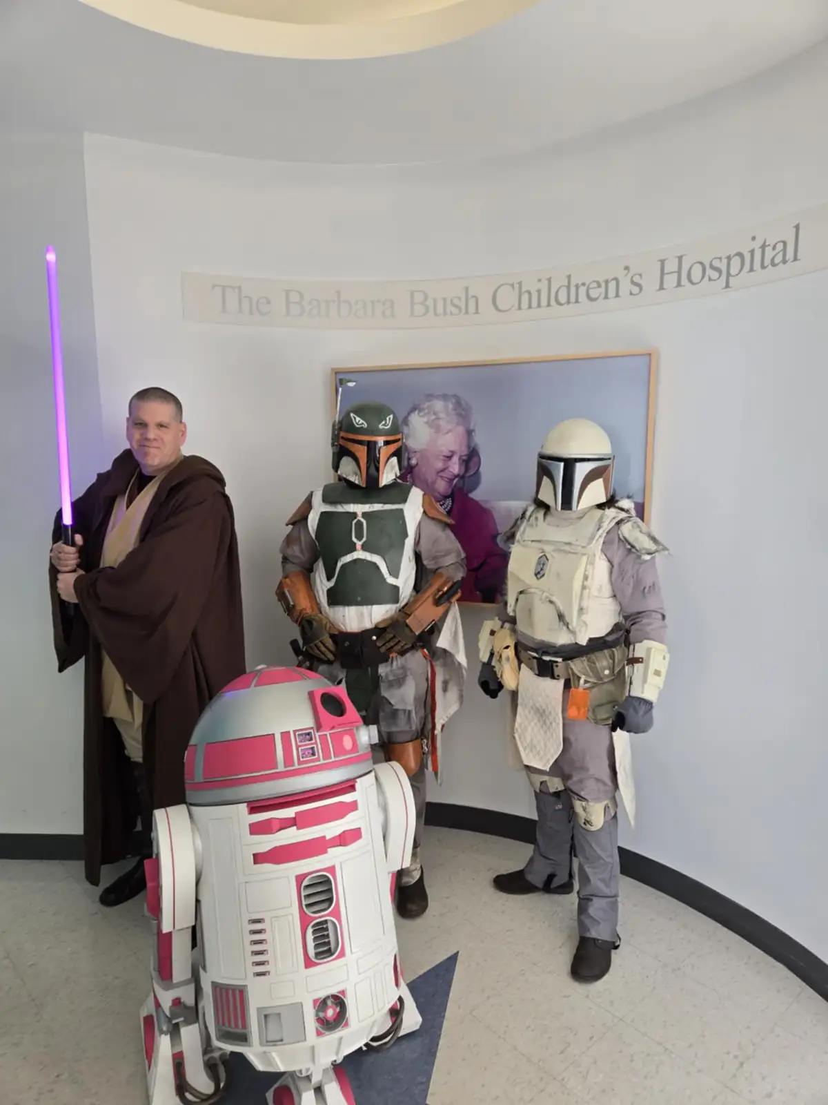 Four people in Star Wars costumes — a Jedi with a purple lightsaber, Boba Fett, a Mandalorian, and a pink-and-white R2-D2…
