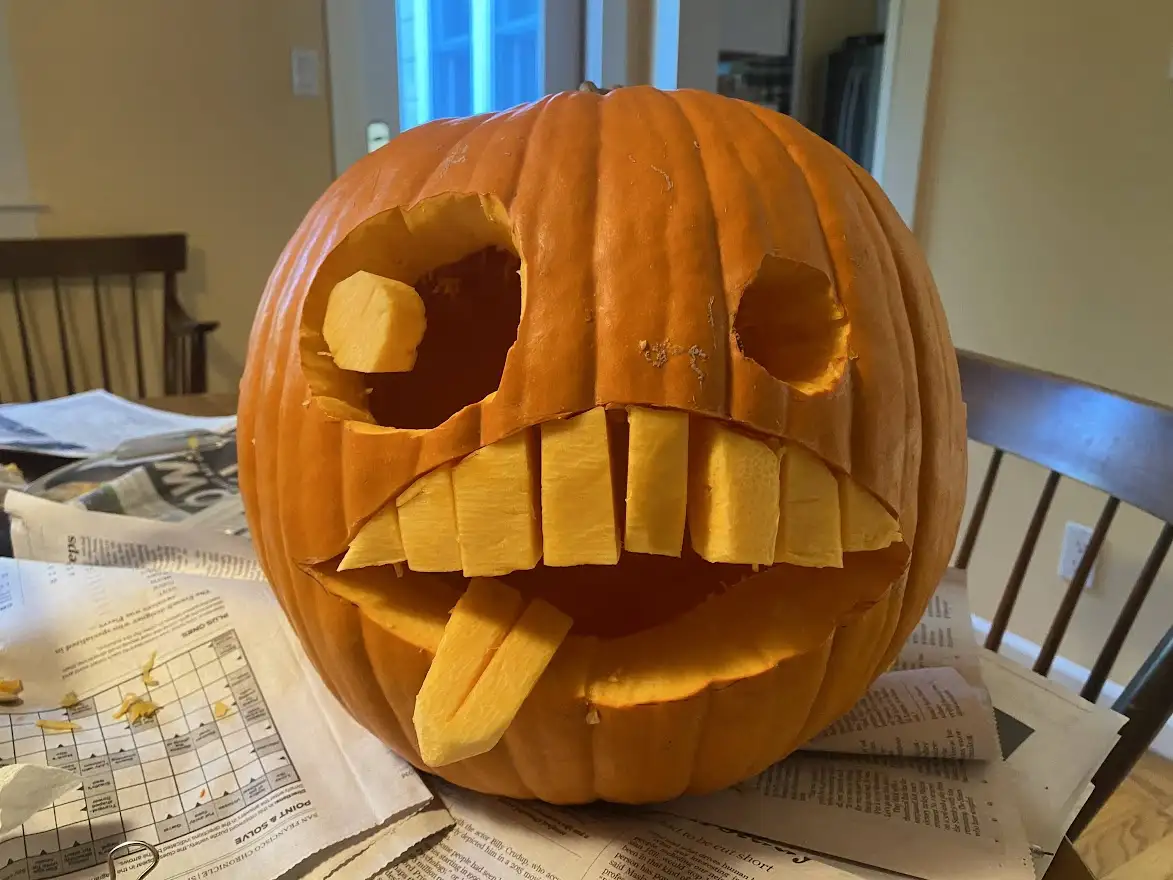 A partially carved orange pumpkin sits on newspaper-covered furniture, its eyes and mouth cut out with uneven, blocky teeth…