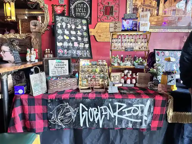 A merchandise table covered in a red-and-black plaid cloth displays various fantasy-themed items like stickers, bottled…