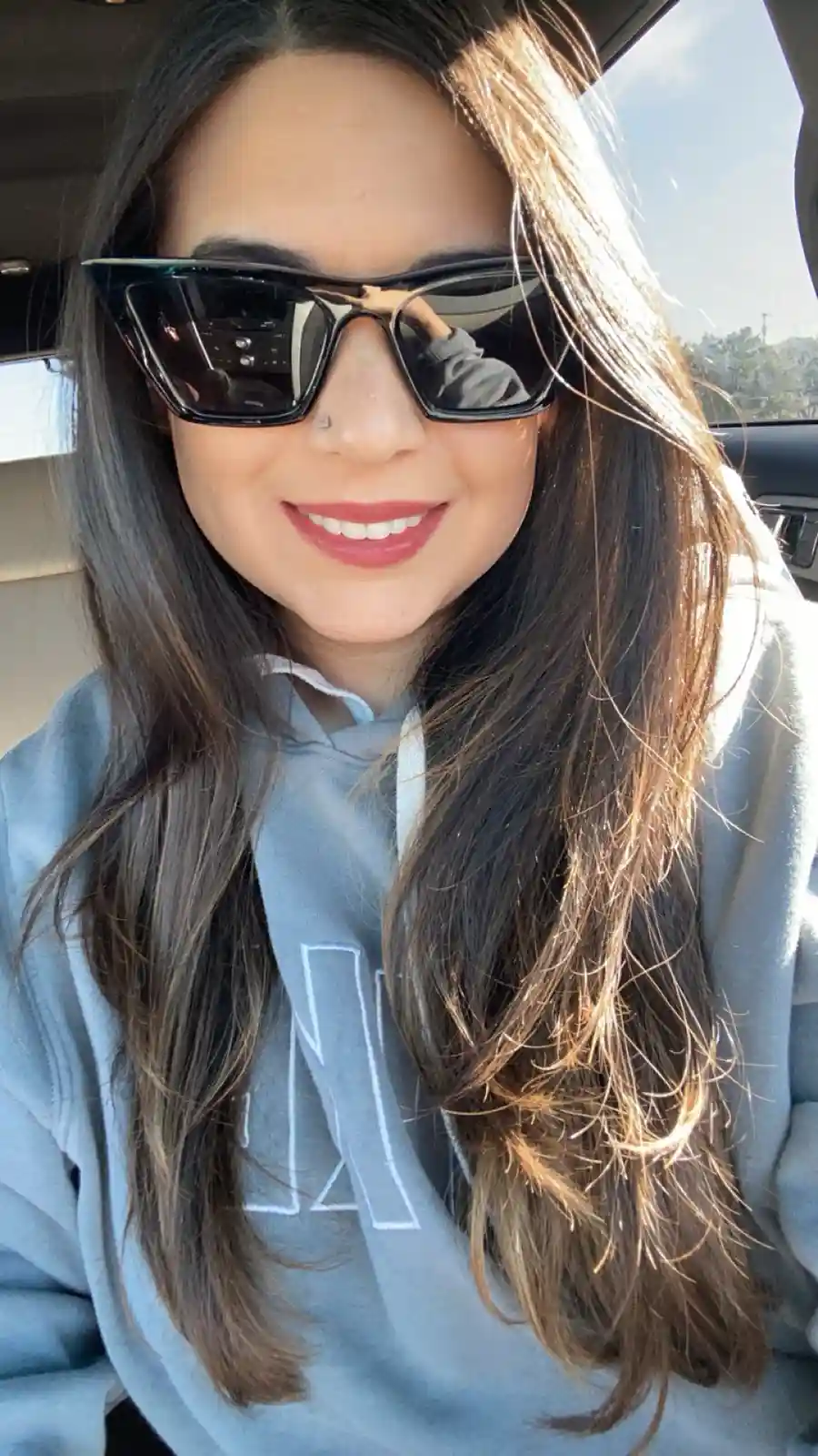A woman with long dark hair and large black sunglasses smiles while sitting in a car, wearing a light blue hoodie with a…