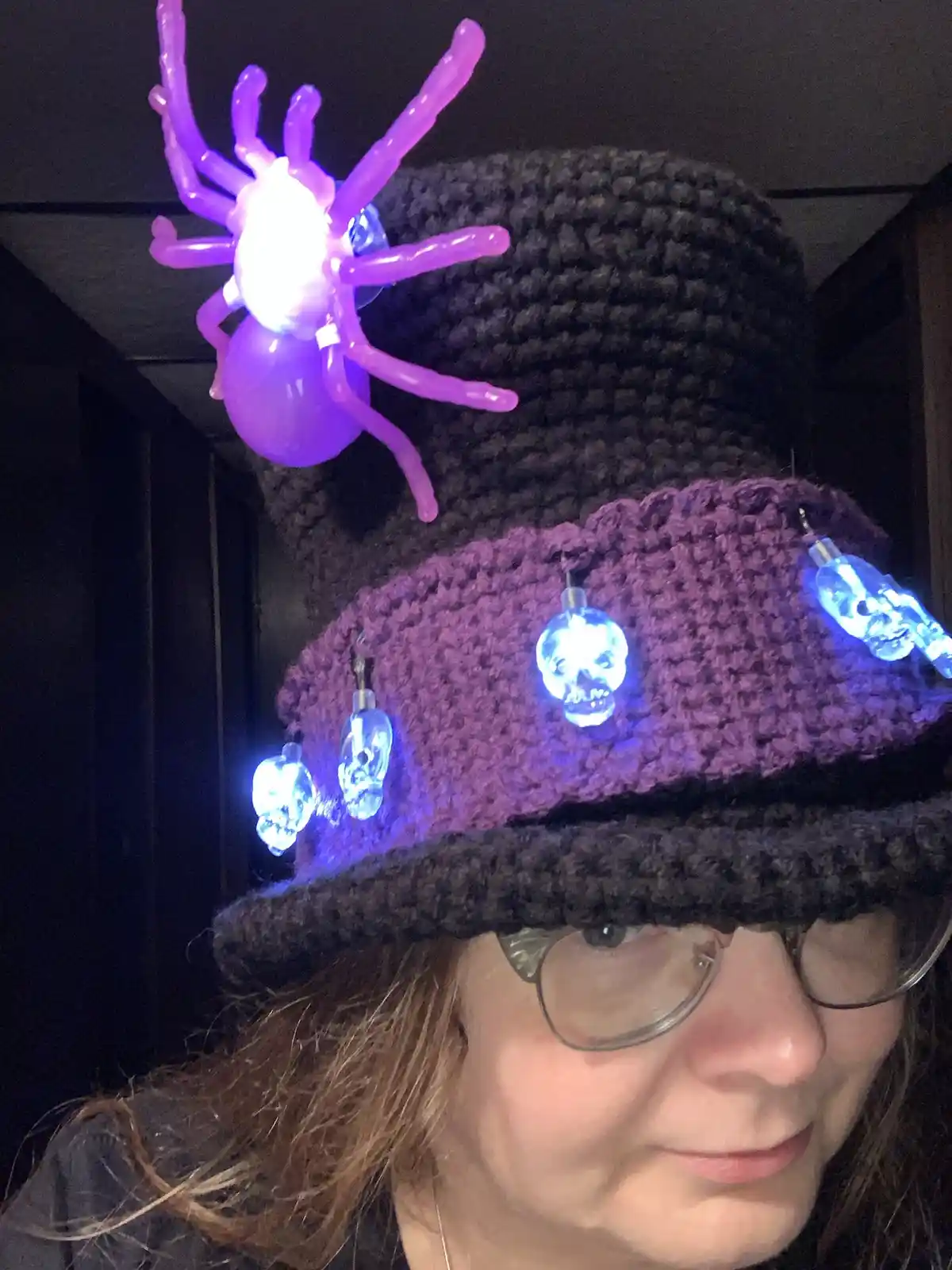 A person wearing a crocheted witch hat adorned with glowing skull lights along the brim and a large, illuminated purple…