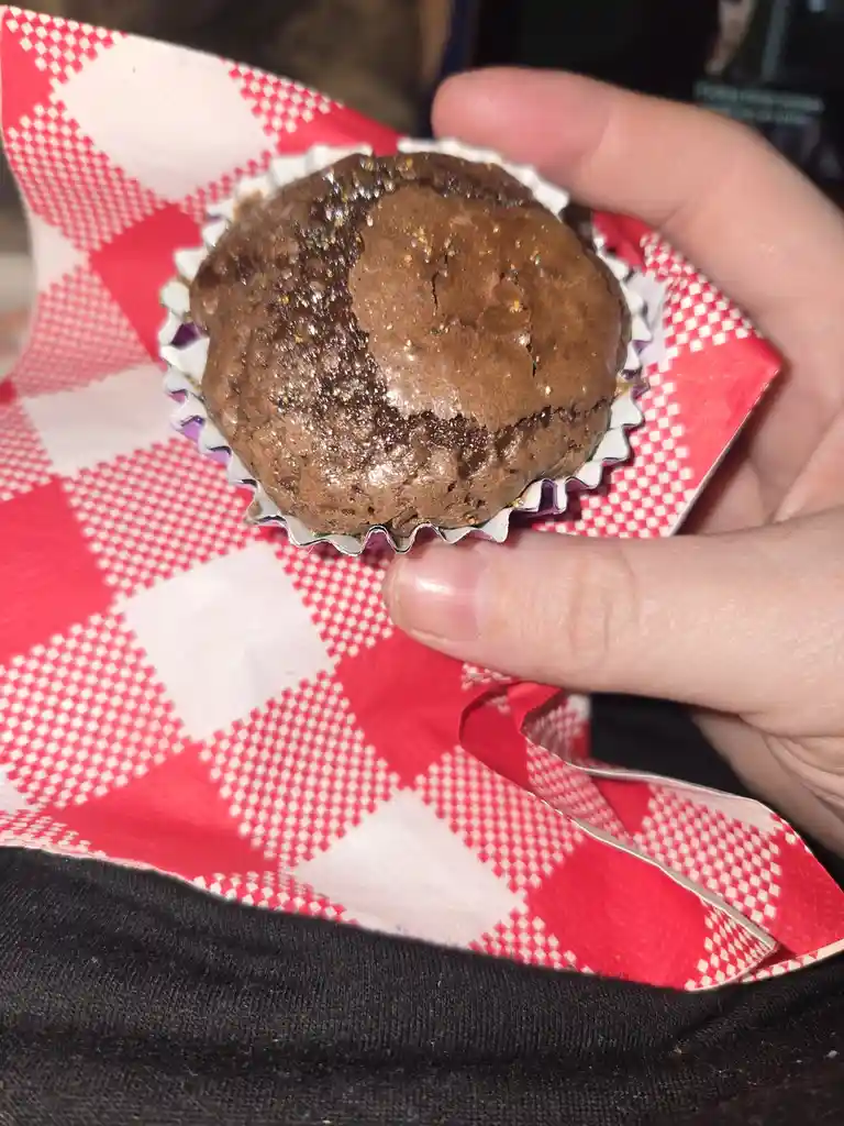 A hand holds a chocolate muffin in a white paper liner, resting on a red and white checkered paper. The muffin's top is…