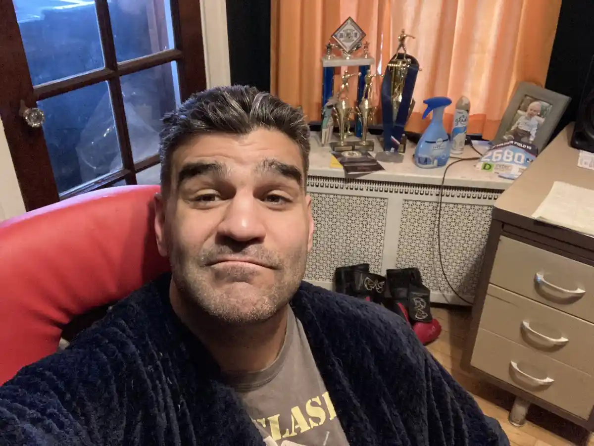 A man with graying hair and a beard, wearing a dark robe over a t-shirt, sits in a red chair and stares at the camera with a…