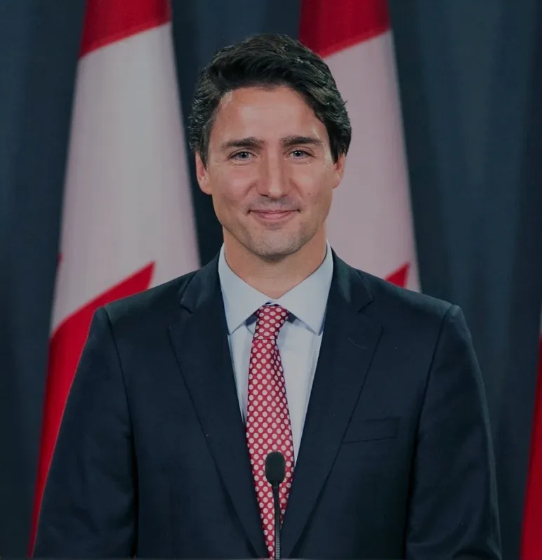 A man in a dark suit and light blue shirt stands at a podium with a microphone, wearing a red tie with white polka dots…