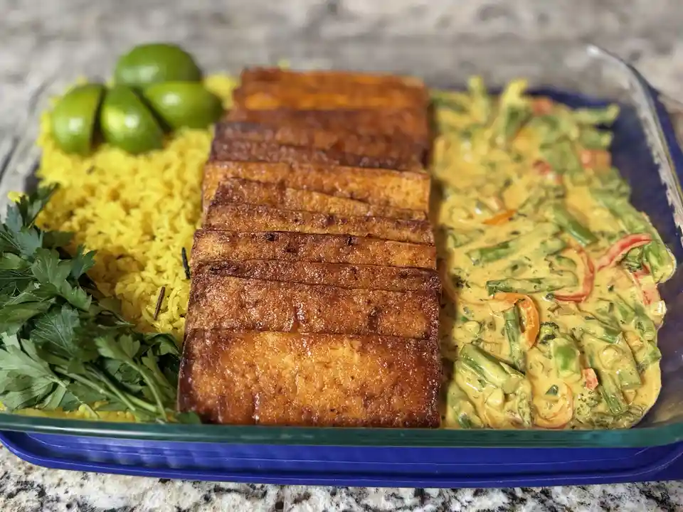 A glass dish contains a meal with yellow rice, sliced tofu, and a creamy vegetable curry, garnished with fresh parsley and…