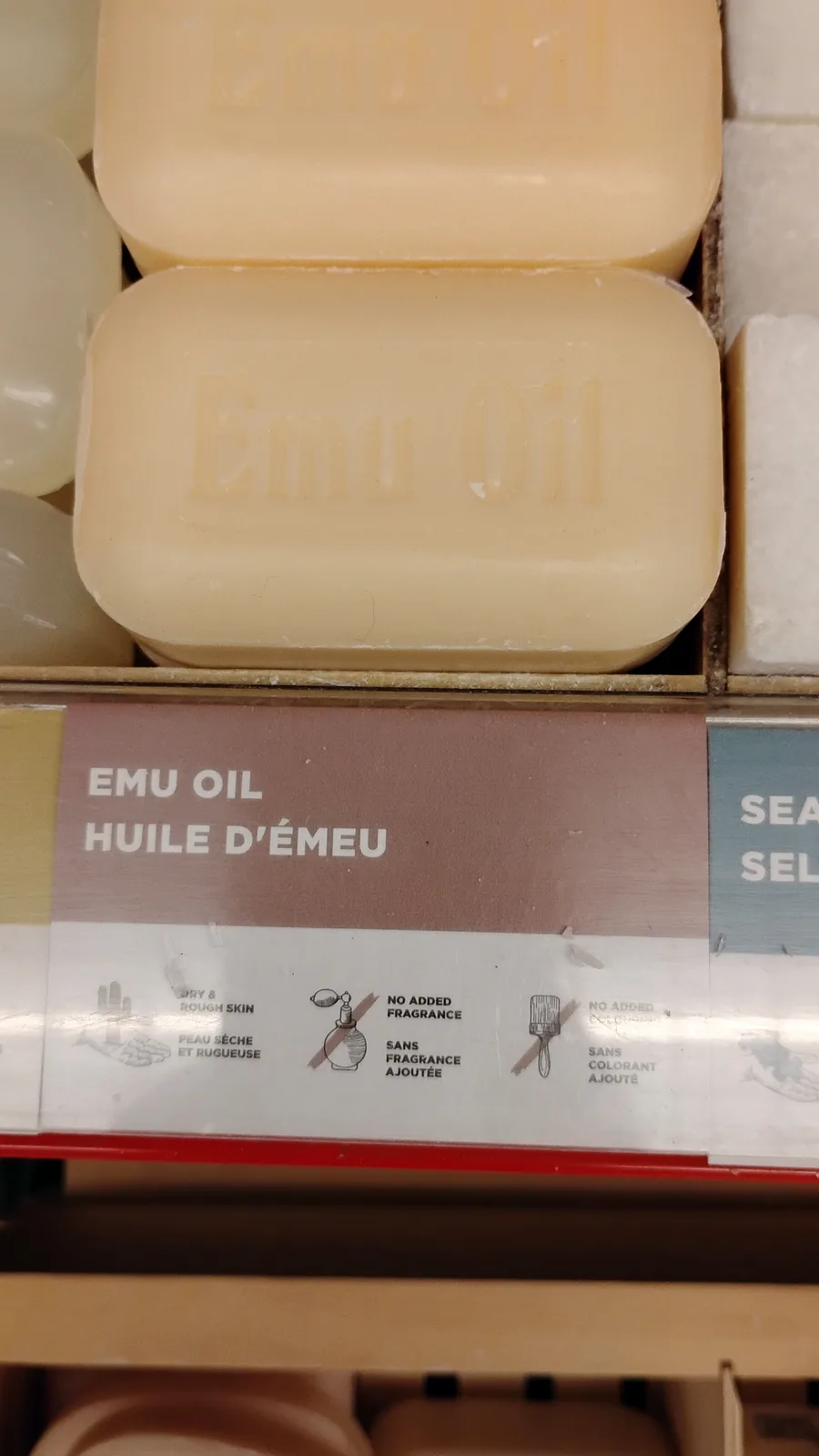 A close-up photo of beige rectangular soap bars labeled 'Emu Oil' on a store shelf, with a product label below stating 'EMU…