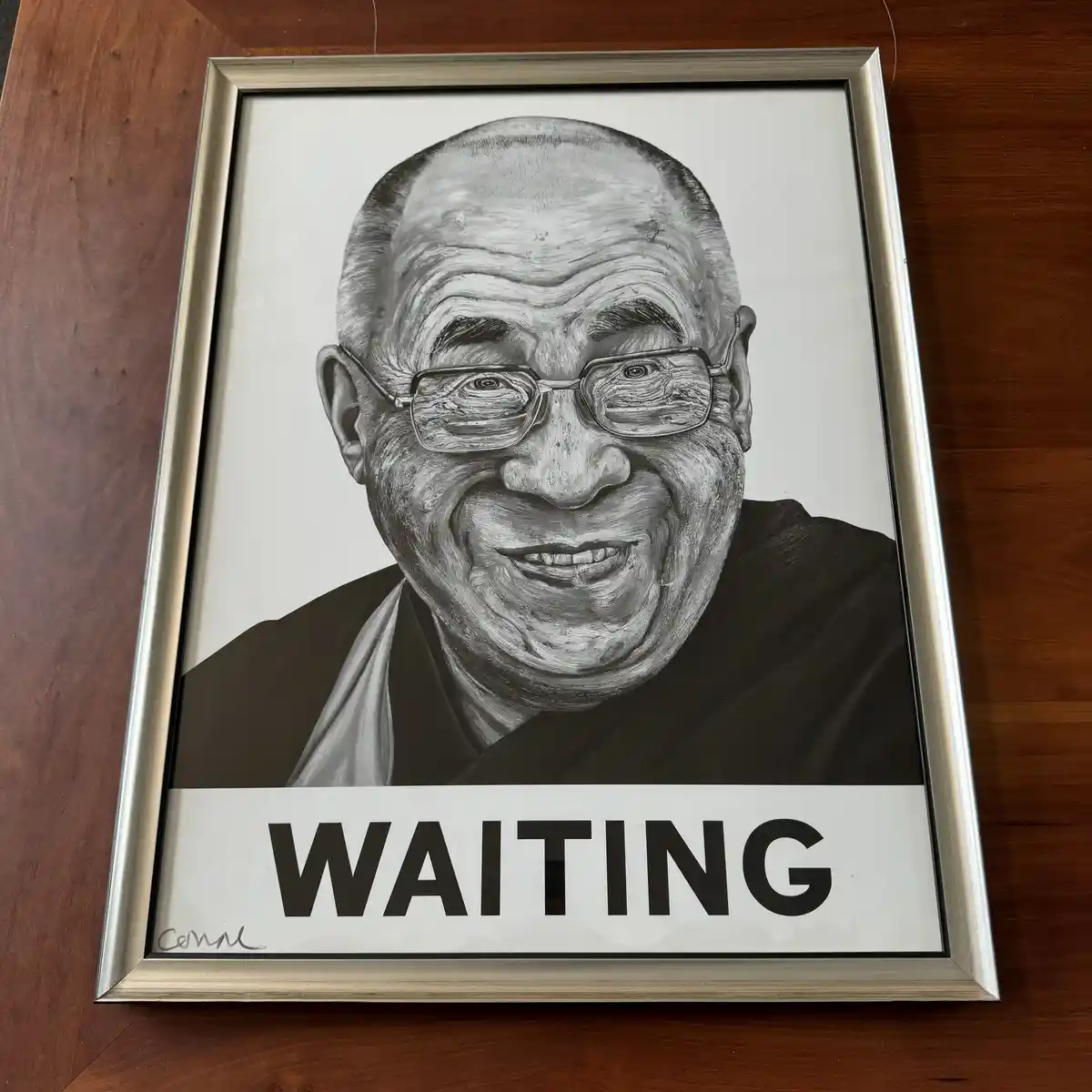 A framed black-and-white portrait of the Dalai Lama, smiling warmly while wearing glasses and traditional robes, has the…