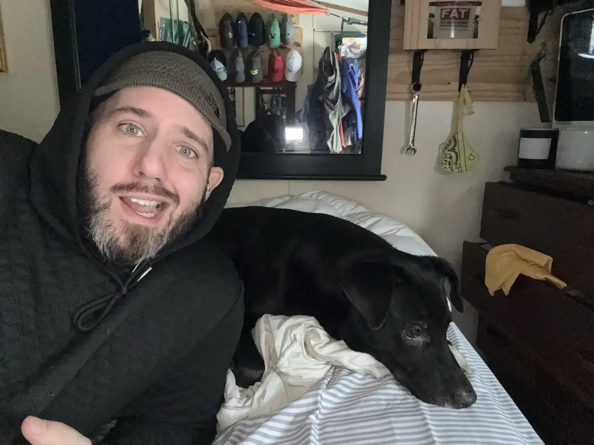 A man in a black hoodie and beanie smiles at the camera while lying on a bed next to a black dog that is resting its head on…