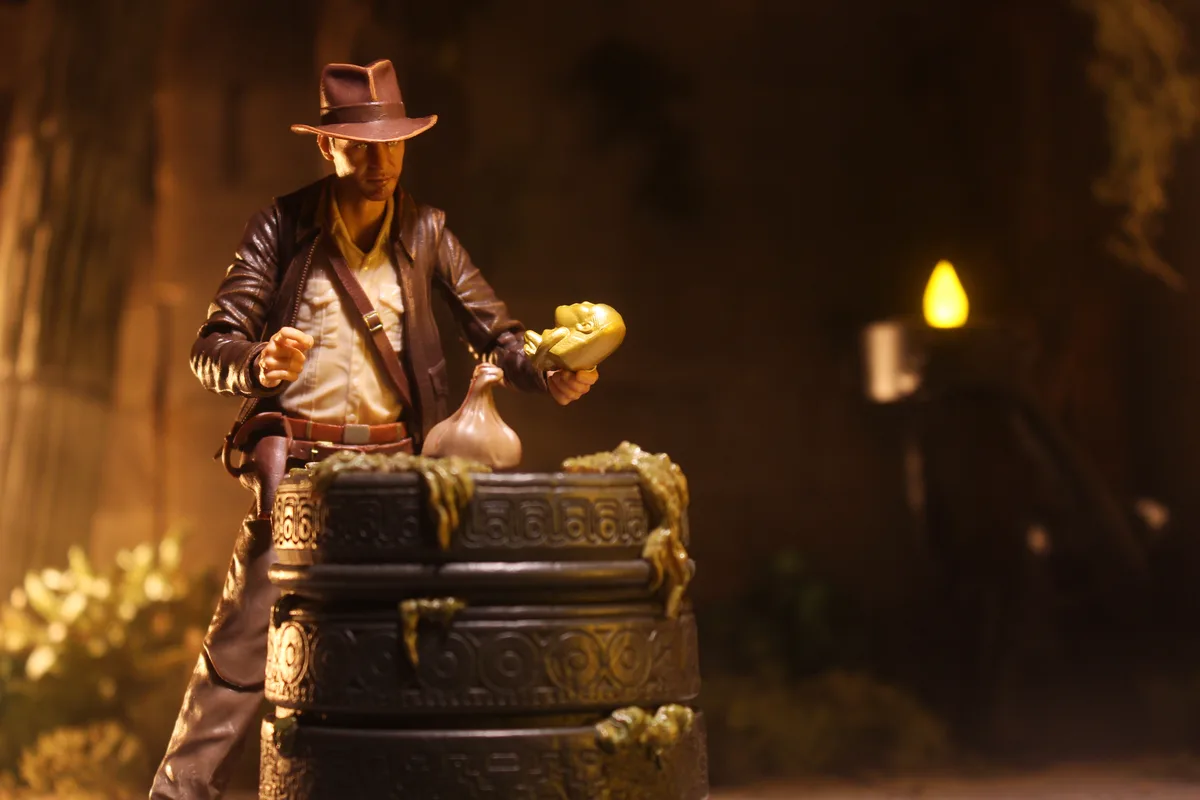 A figurine of Indiana Jones stands in a dimly lit, jungle-like setting, holding a golden idol in one hand and a garlic bulb…