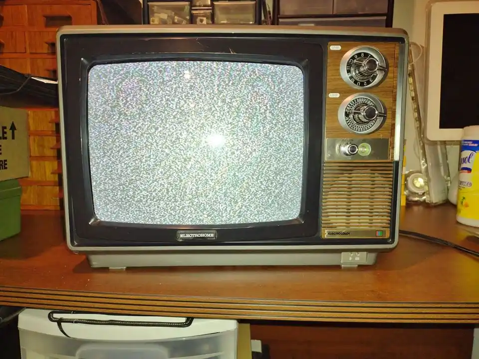 A vintage Electrohome television with a wooden side panel and two dials sits on a wooden desk, its screen filled with static…