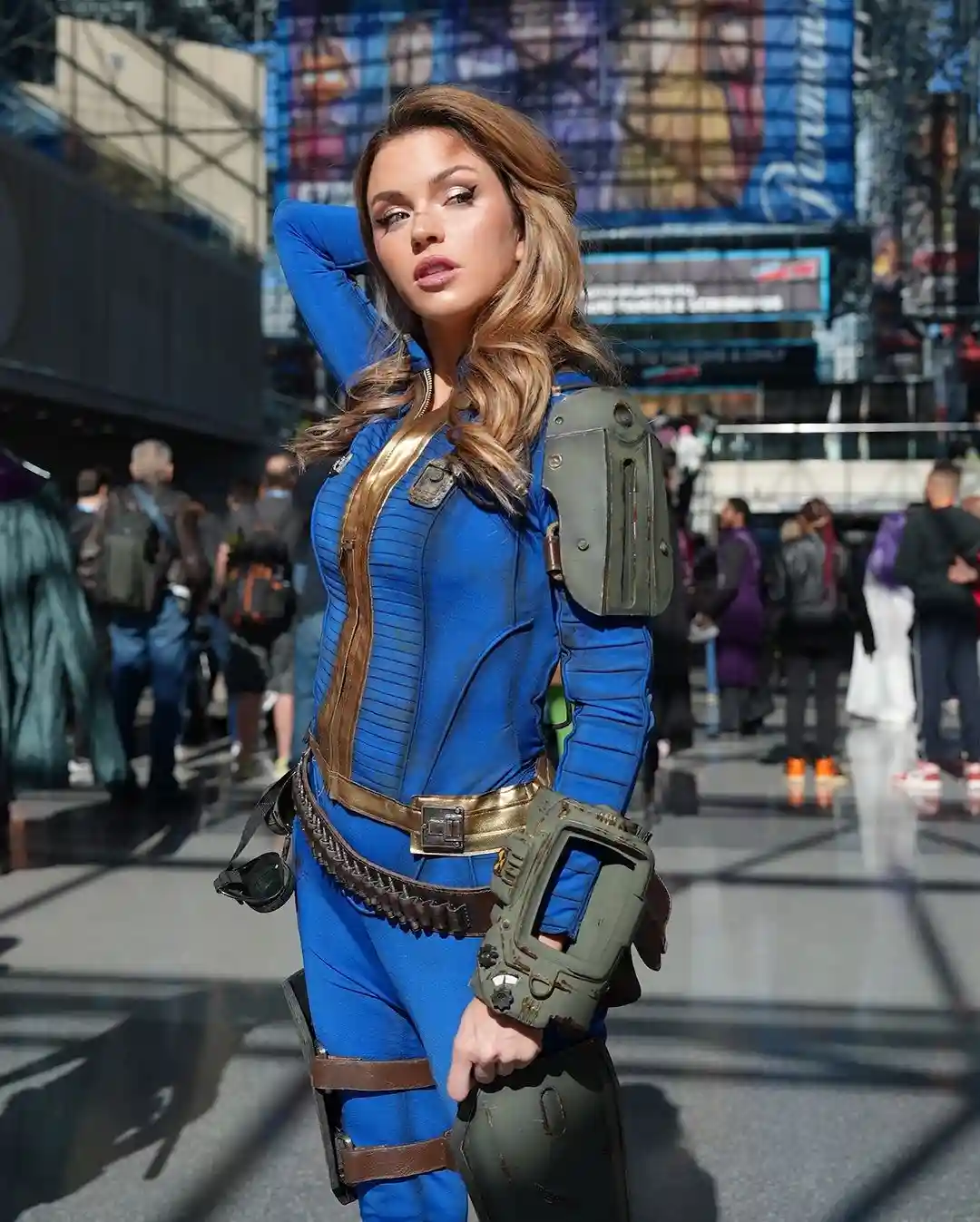 A woman in a detailed blue and gold Fallout-style cosplay stands confidently in a busy urban plaza, one hand on her head…