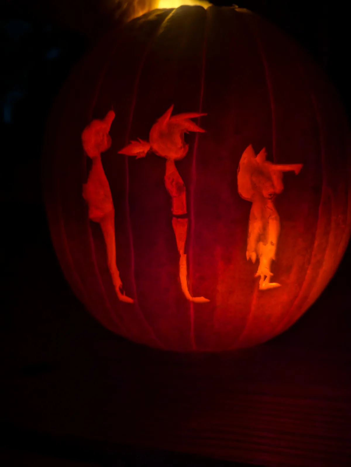 A carved pumpkin illuminated from within, showing three stylized, cartoonish figures with spiky hair and long limbs…