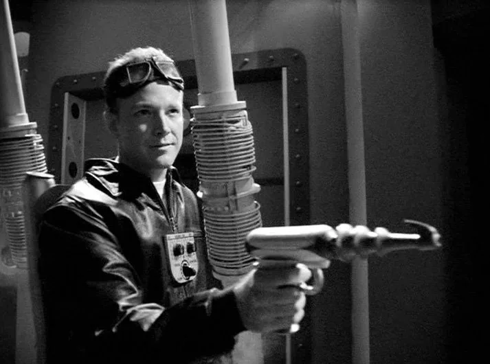 A black-and-white image shows a man in a leather flight jacket with goggles pushed up on his head, holding a…