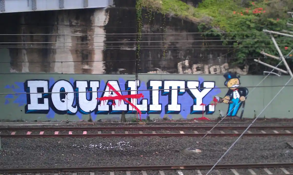 A graffiti mural on a green wall beside train tracks shows the word 'EQUALITY' in large white letters with a red…
