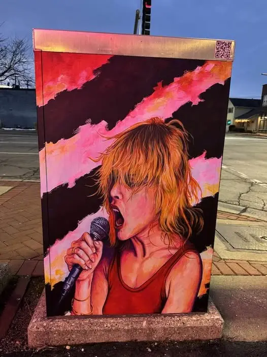 A painted mural on a utility box depicts a woman with messy blonde hair, wearing a red tank top, singing passionately into a…