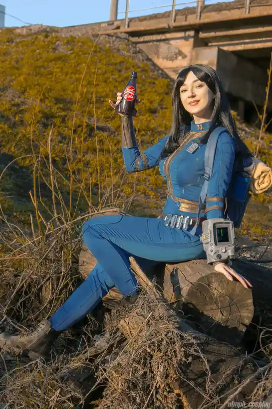 A person in a blue Fallout 4 power armor cosplay sits on a log outdoors, holding up a bottle of Nuka-Cola. The bottle has a…