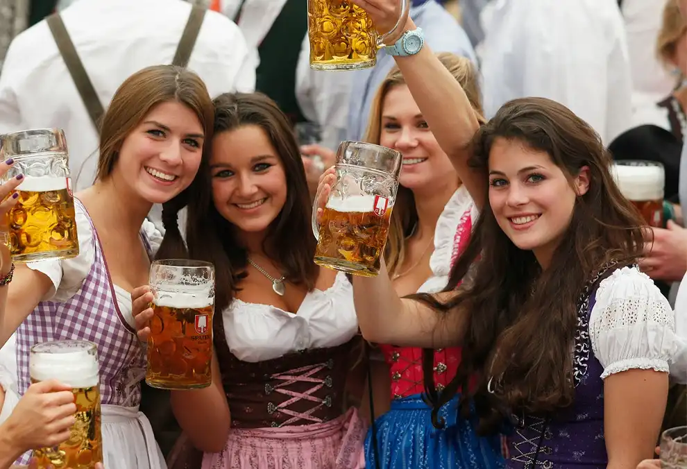 four smiling young women in traditional dirndls hold large steins of beer at what appears to be a German festival. One woman…