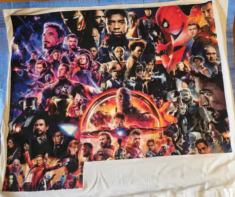 A white towel or blanket with a large, colorful print of the Avengers Endgame movie poster, featuring dozens of Marvel…