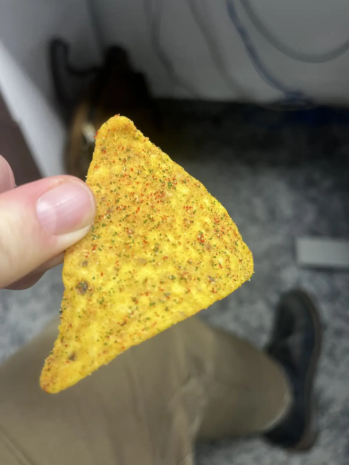 A close-up photo shows a hand holding a single, triangular, yellow corn chip covered in red and green seasoning. The…