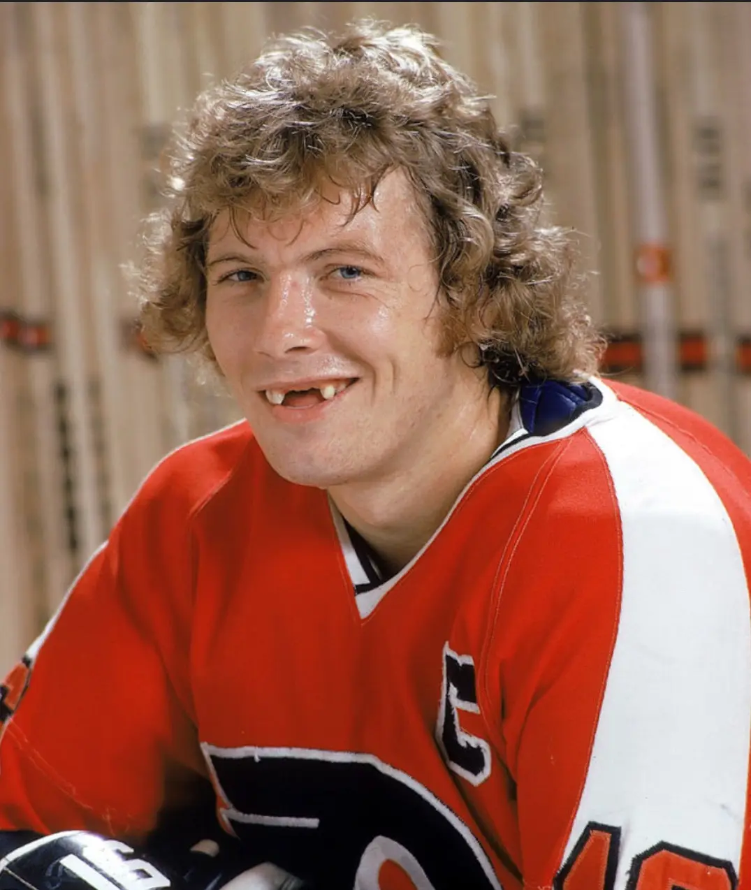 a smiling man in a red and white hockey jersey with visible missing front teeth, set against a background of hockey sticks…