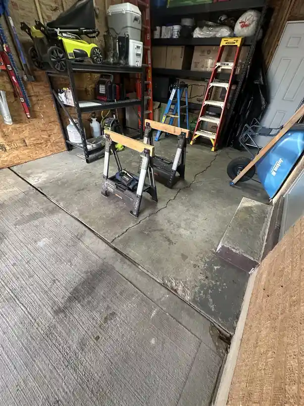 A cluttered garage floor shows a distinct boundary where new gray wood-look laminate flooring meets old cracked concrete…