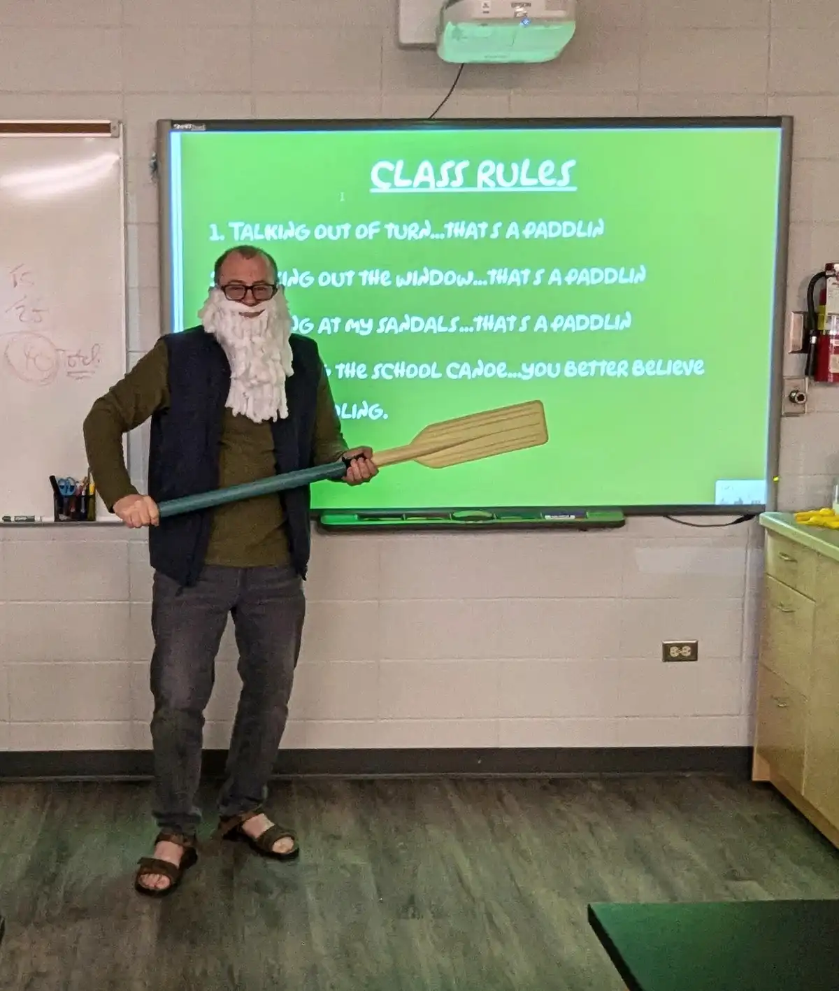 A man wearing glasses and a fake white beard stands in a classroom holding a large paddle, smiling. Behind him, a green…