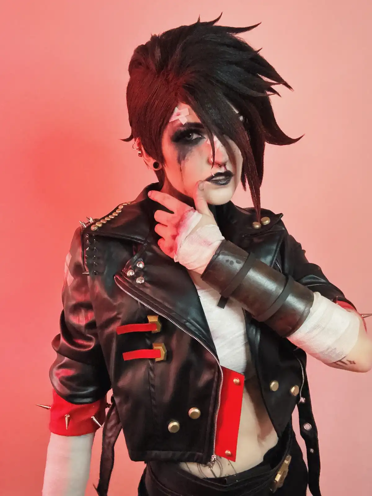A person in a punk-style cosplay with black spiky hair, heavy makeup, and bandages on their face and hands, wearing a black…