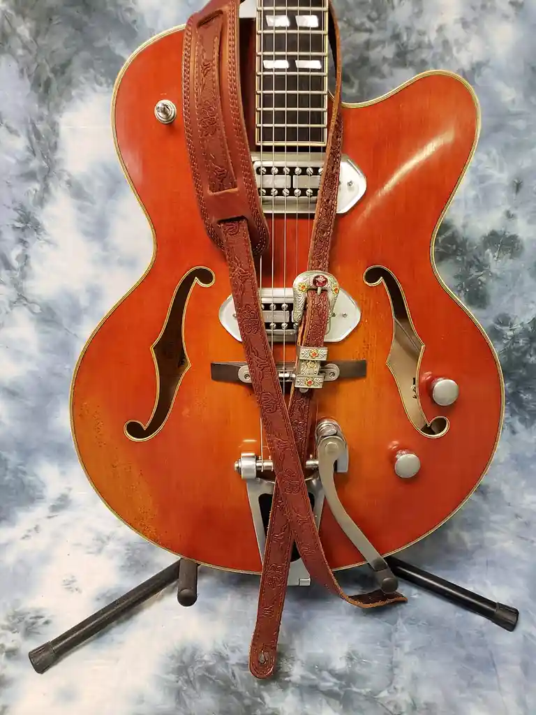 A close-up photo of a reddish-orange archtop electric guitar with a decorative brown leather strap adorned with floral…