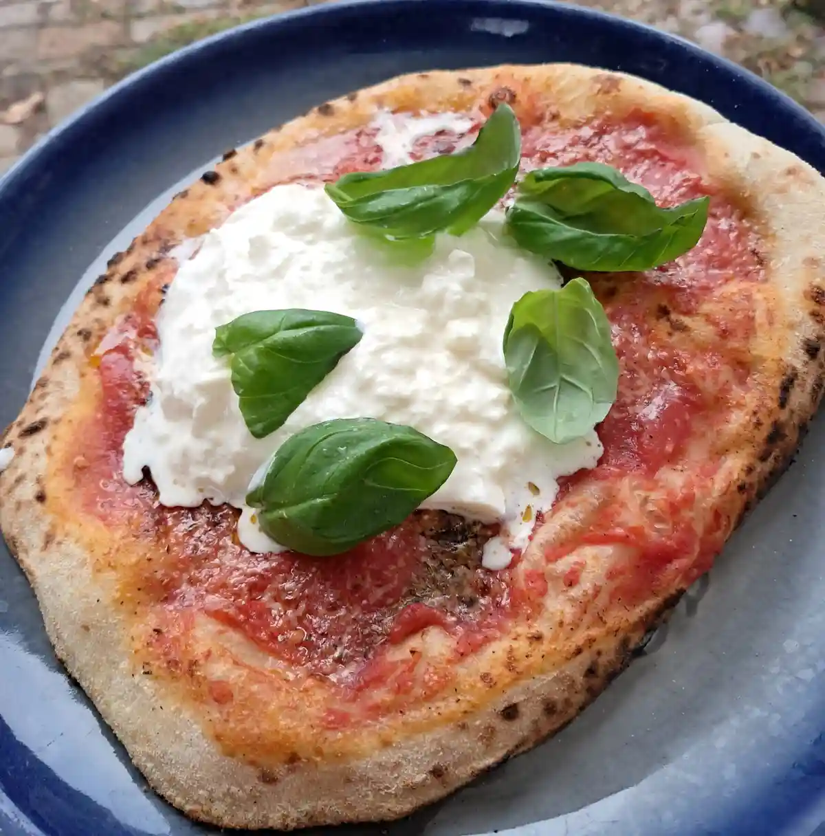 A close-up photo shows a small, rustic pizza on a dark blue plate, featuring a golden-brown crust with charred spots, a…