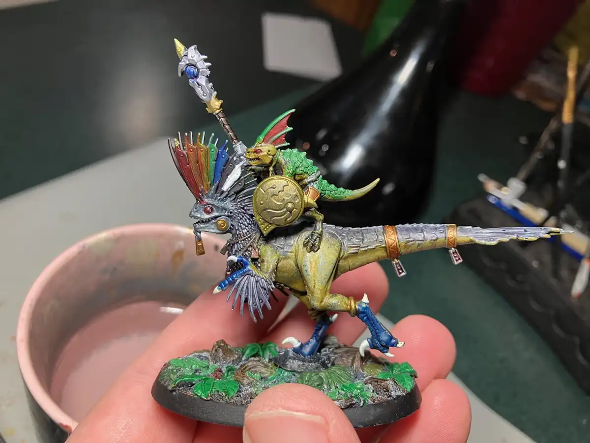 A hand holds a highly detailed, painted miniature of a dragon rider. The rider is a small, green, reptilian creature with…