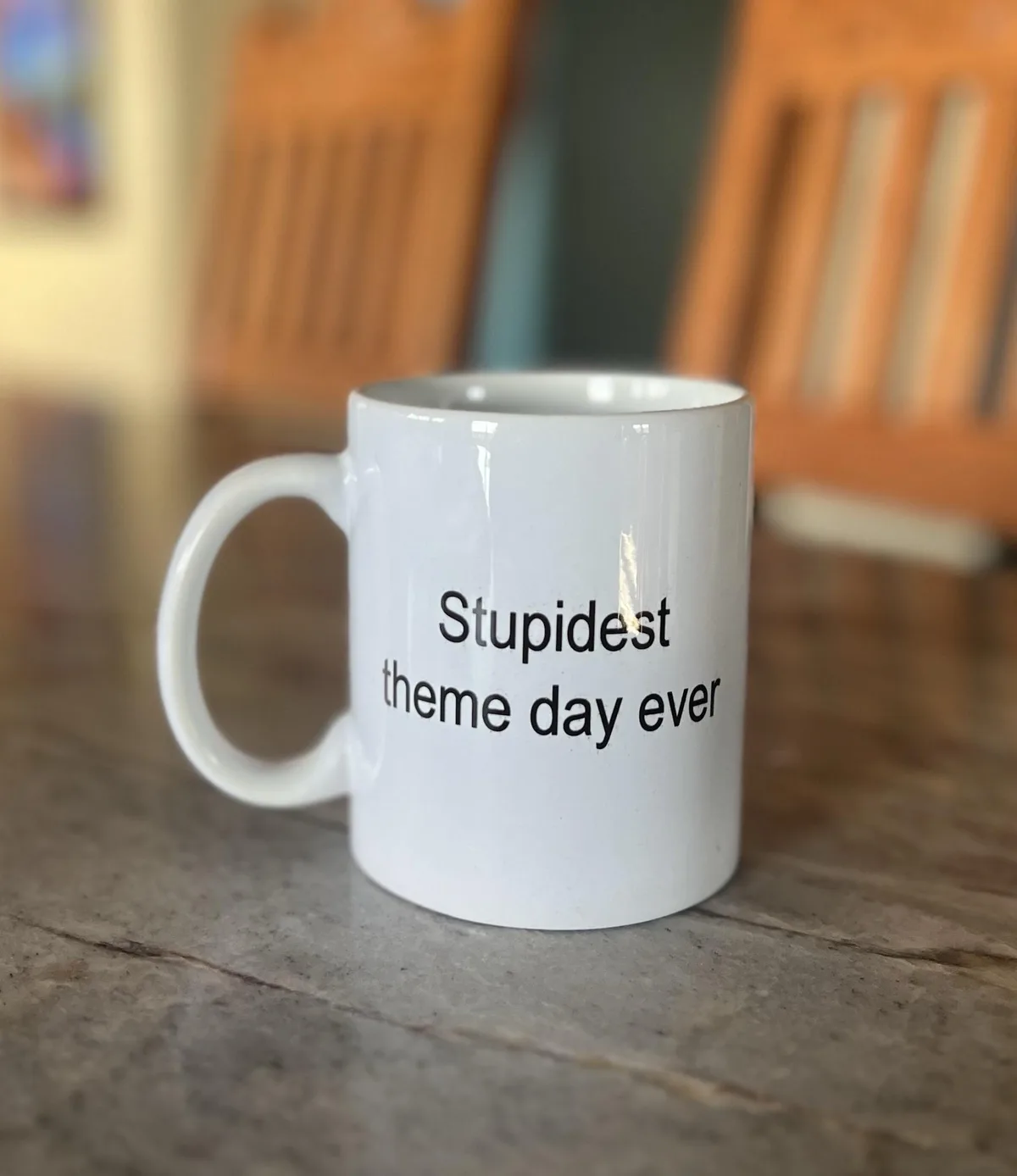 A plain white ceramic mug with black text reading 'Stupidest theme day ever' sits on a wooden table, with blurred wooden…