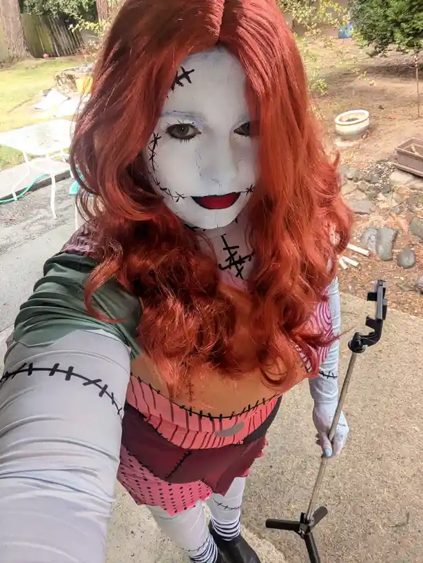 A person with long red hair and white face paint with black stitches takes a selfie outdoors, holding a selfie stick. Their…