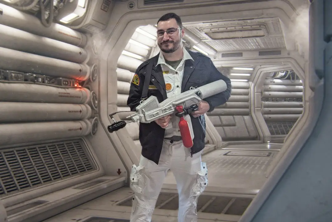 A man in a sci-fi spaceship corridor, wearing a jacket and glasses, holds a large, silver, futuristic-looking weapon with a…