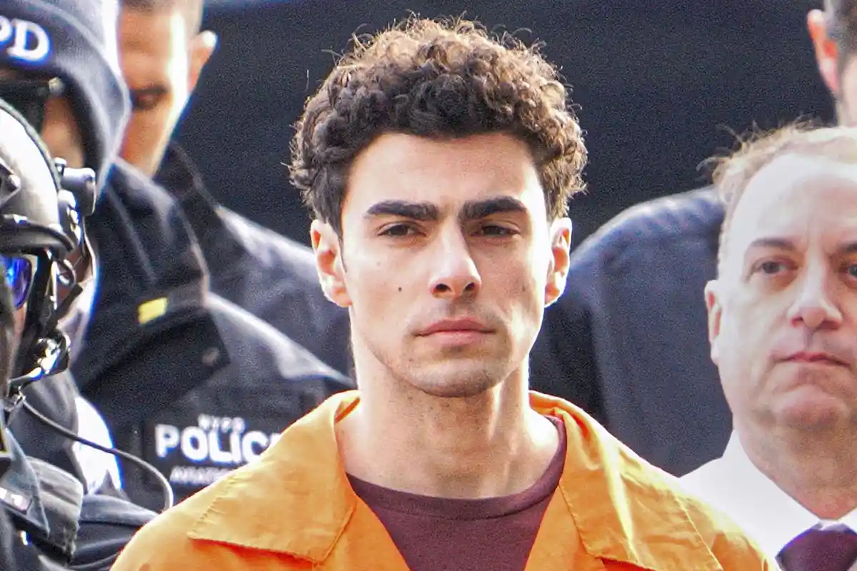 A man with curly dark hair wearing an orange prison jumpsuit is flanked by NYPD officers and another man in a suit. The…