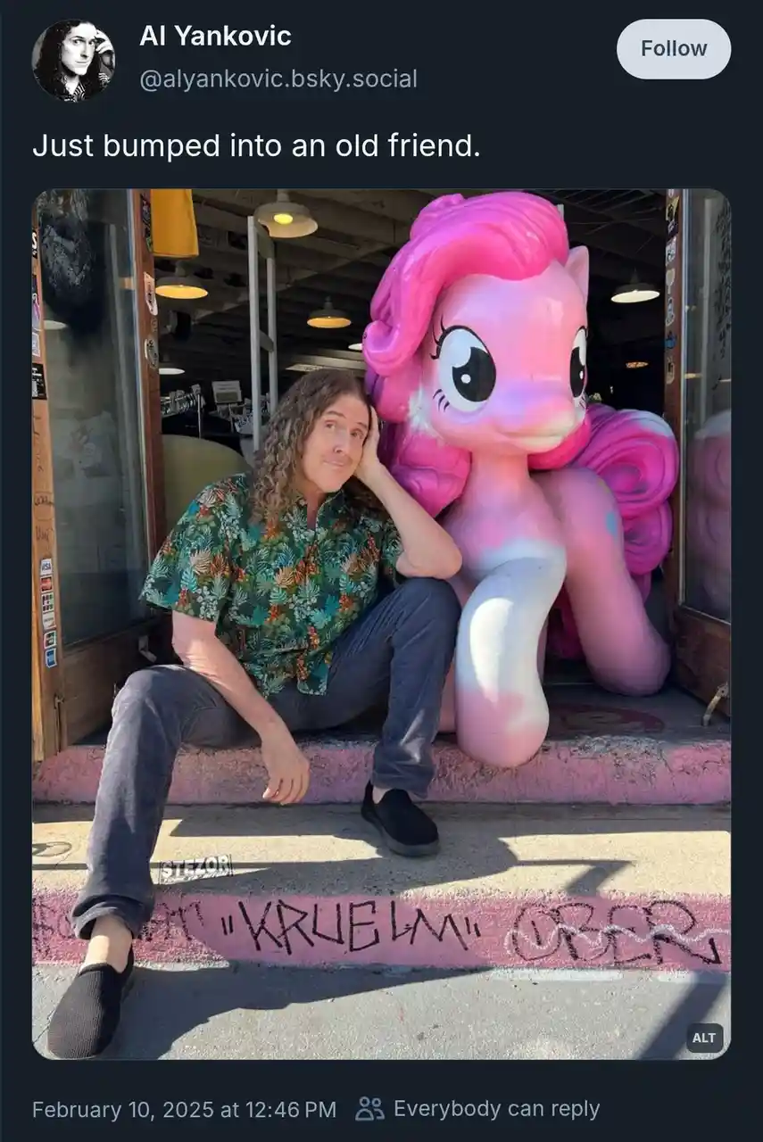 A man with long curly hair, wearing a patterned shirt and dark pants, sits on a pink-painted curb next to a large, pink…