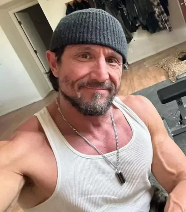 A middle-aged man with a beard and a grey beanie smiles at the camera while wearing a white tank top and a dog tag necklace…