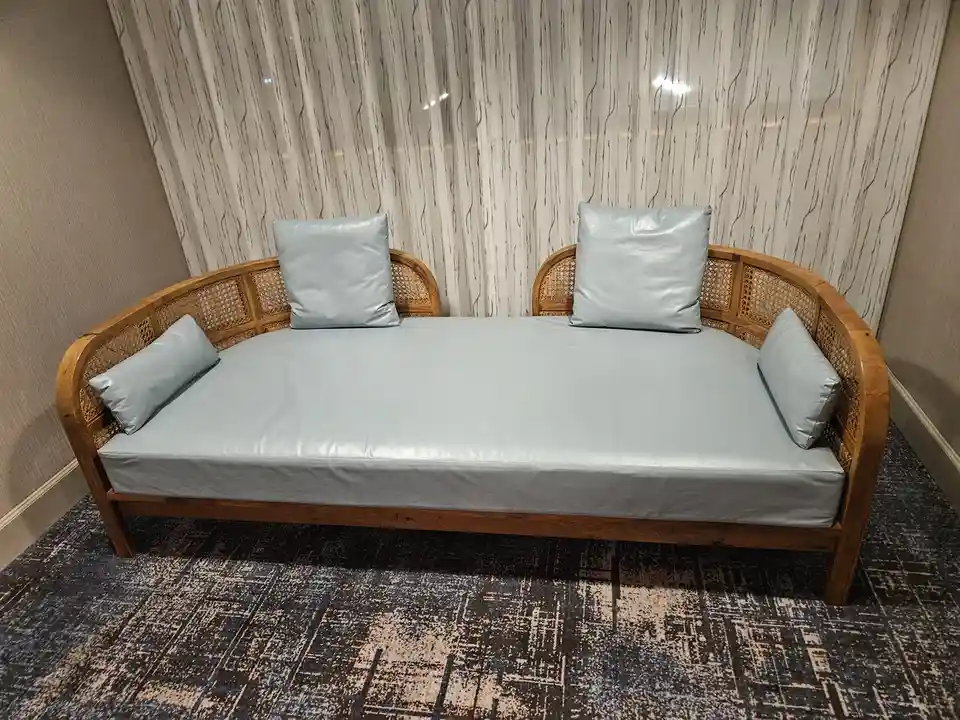 A light blue daybed with a wooden rattan frame and four matching pillows sits in a room with patterned carpet and sheer…