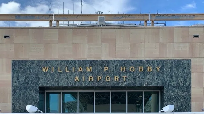 A photograph of the exterior of William P. Hobby Airport, showing its name engraved in gold letters on a dark marble panel…