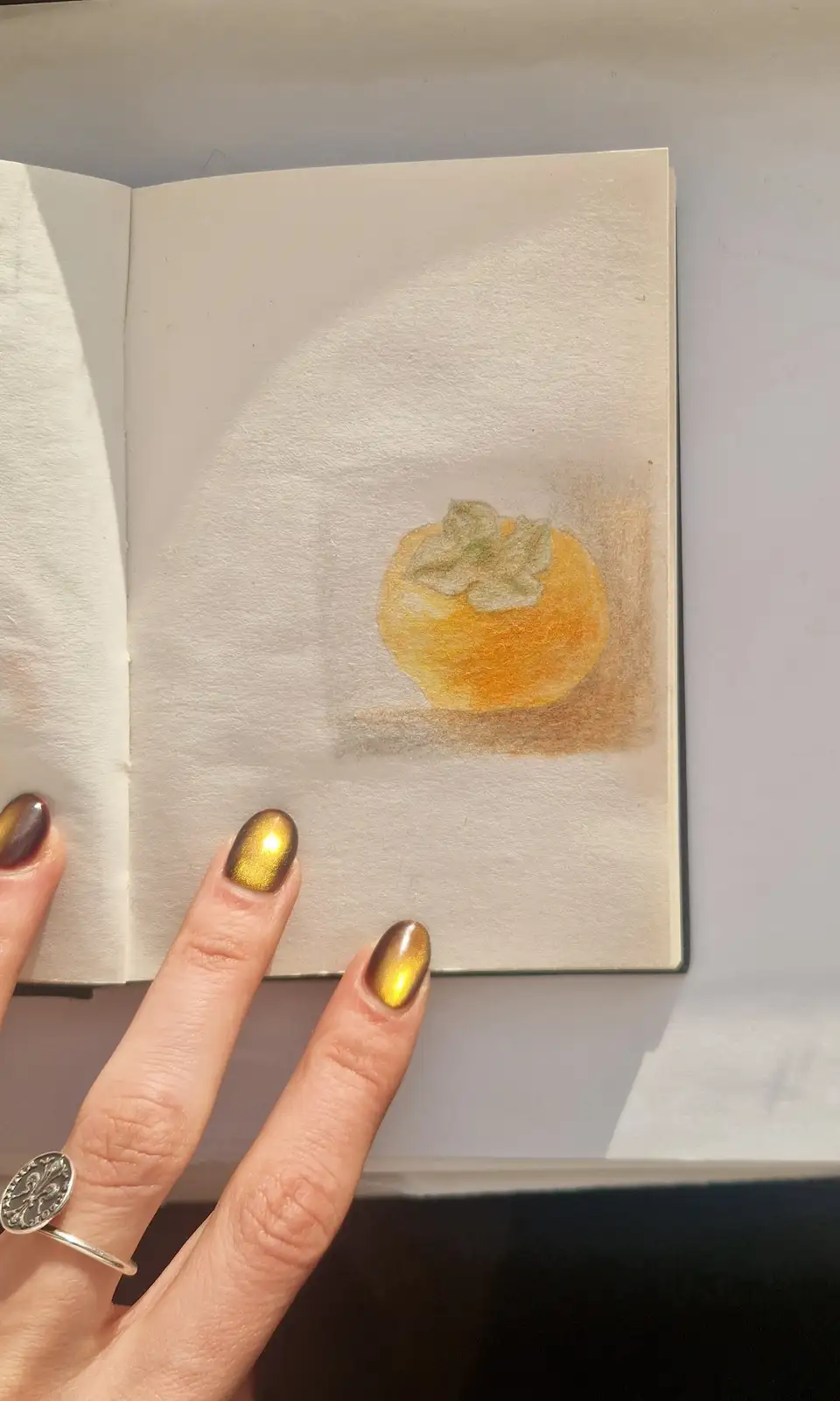 A hand with gold and brown metallic nail polish holds open a small sketchbook displaying a colored pencil drawing of a…