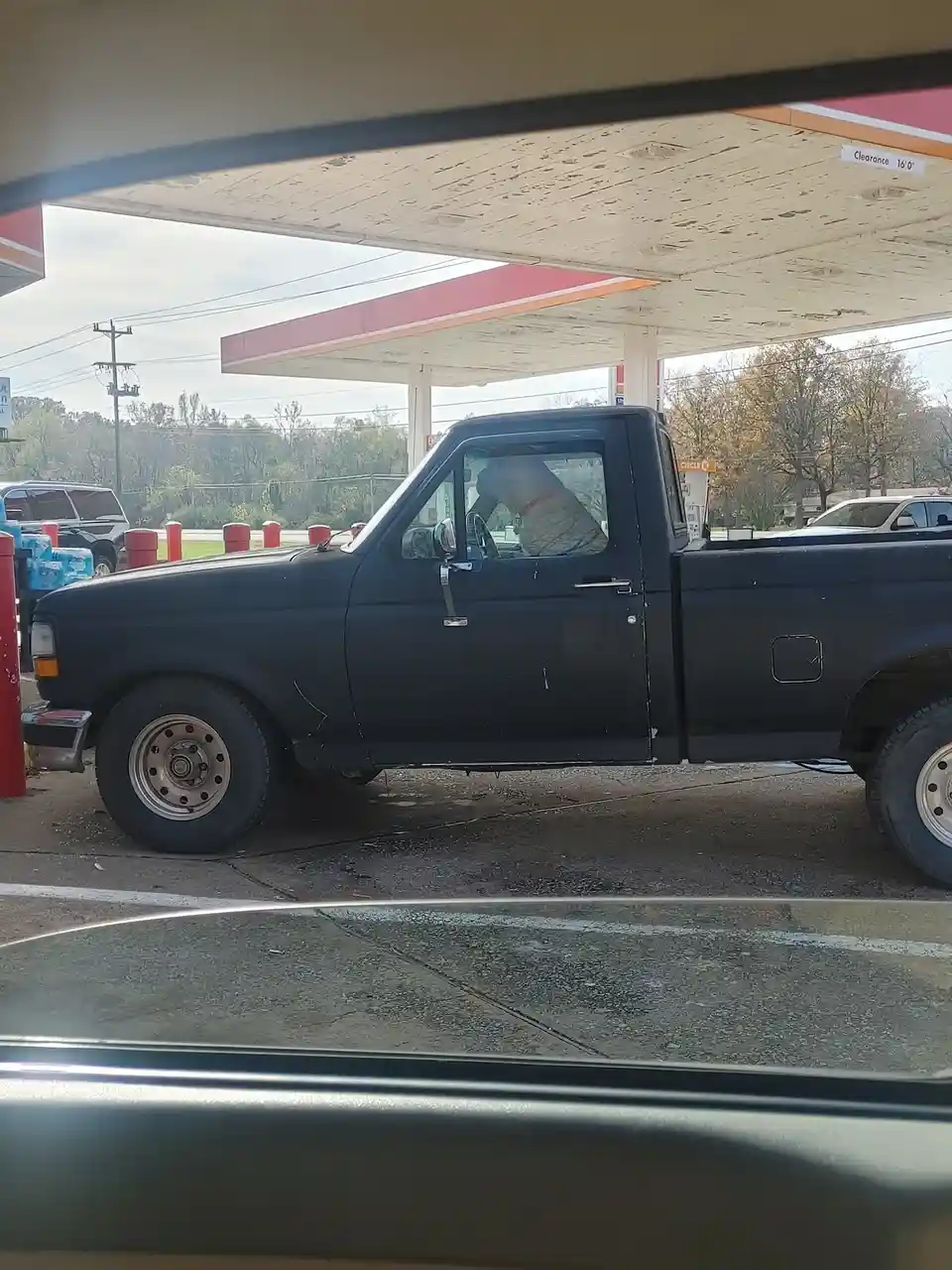 A black pickup truck is parked at a gas station, and a large, light-colored dog is sitting in the driver's seat, looking out…