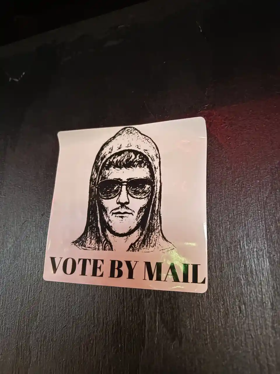 A square sticker with a black-and-white illustration of a man wearing a hood and sunglasses, placed on a dark wooden…
