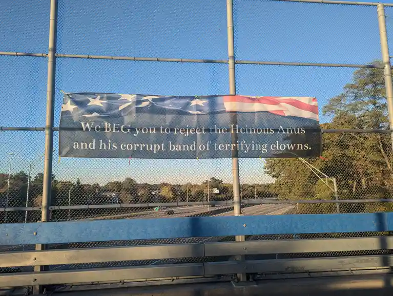 A banner with an American flag design is hung on a chain-link fence overlooking a highway. The banner displays the text 'We…