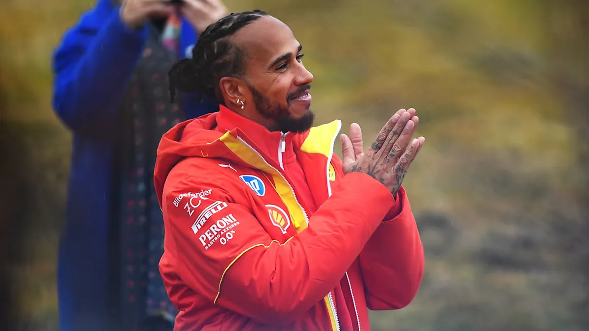 a man in a red and yellow jacket with sponsor logos like Pirelli and Shell, clapping with tattoos visible on his hands…